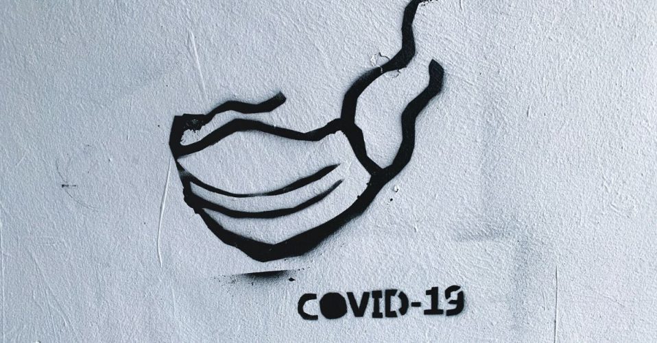 image of a spray painted mask on a white brick wall with spray painted text which says covid-19